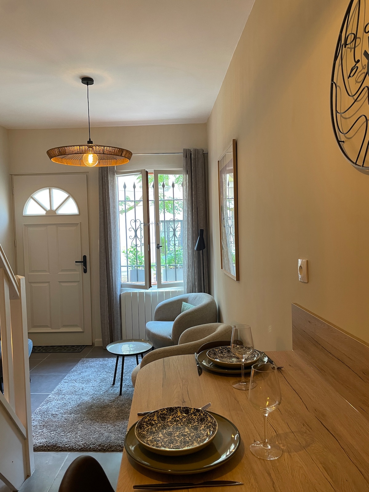 A well-lit dining area features a wooden table set for two, complete with decorative plates and glasses. Soft gray armchairs are positioned nearby, with a window allowing natural light to illuminate the space. A stylish light fixture hangs overhead, enhancing the inviting atmosphere.