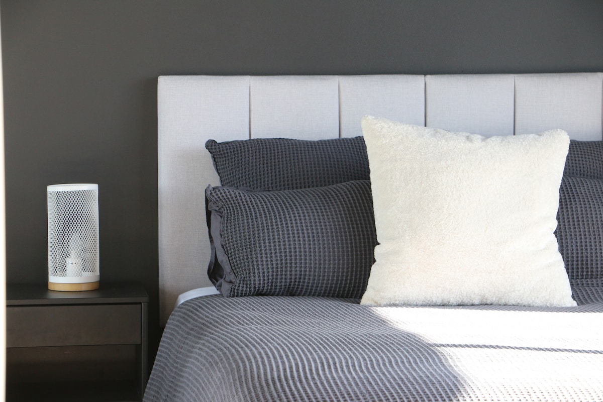 A modern bedroom features a queen-sized bed with a soft grey bedspread and an assortment of patterned pillows. A plush white decorative pillow is placed at the front. A stylish bedside table holds a small, contemporary lamp, reflecting a blend of comfort and elegance.