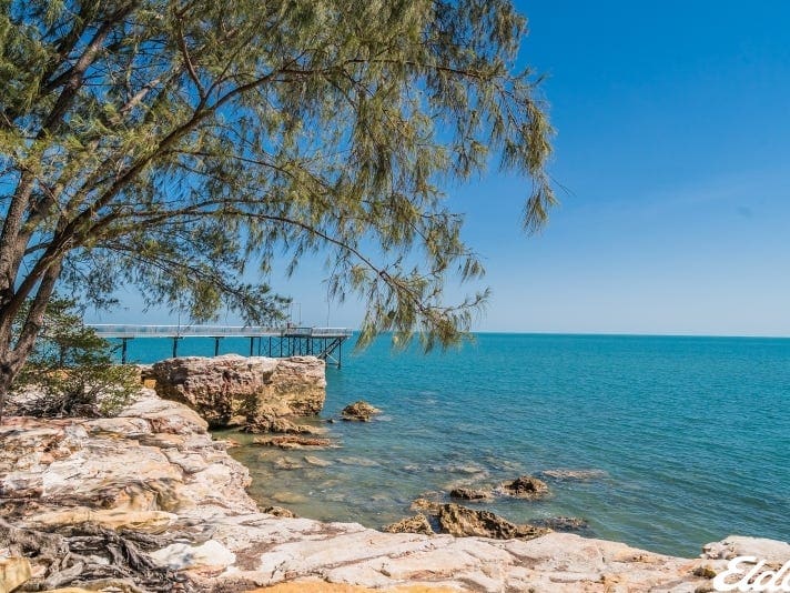 The image illustrates a rocky shoreline with clear blue waters extending into the horizon. A pier is visible in the distance, surrounded by trees with green foliage. The sky is bright, enhancing the tranquil seaside atmosphere.
