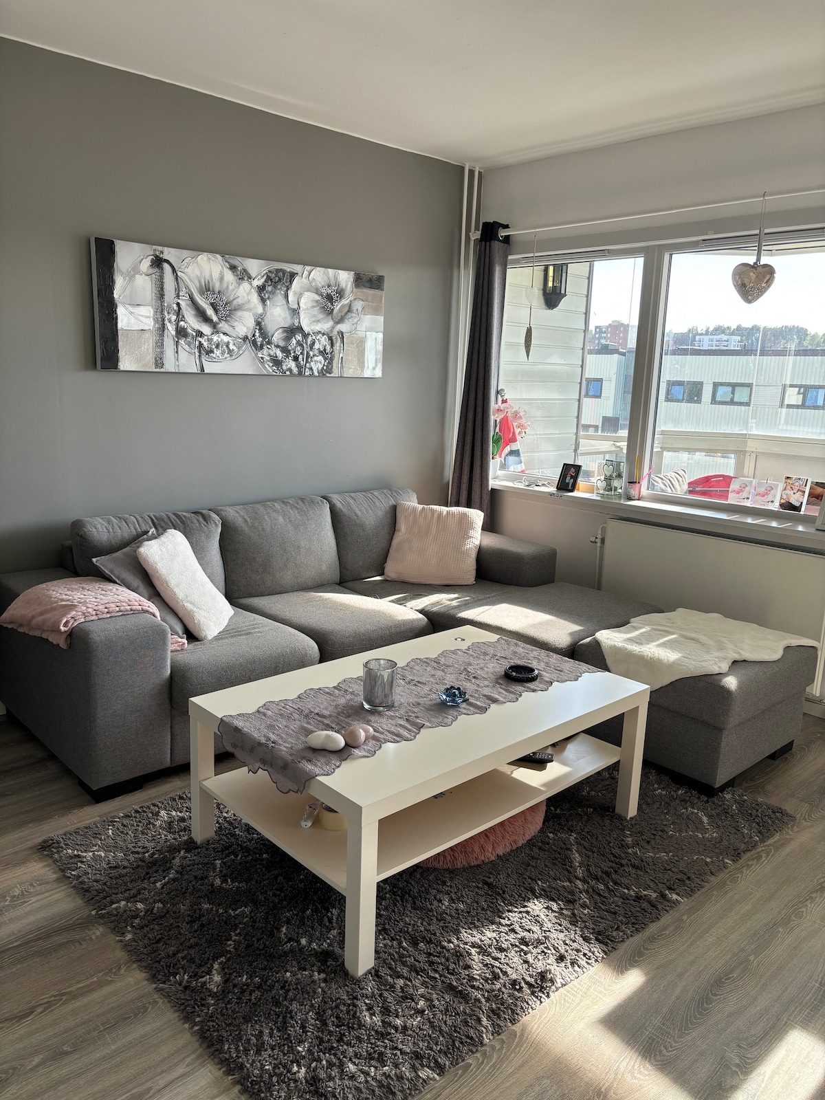 A comfortable living area is presented with a modern gray sectional sofa and a light coffee table. Natural light enters through a large window, highlighting decorative items on the table. A soft rug covers the floor, adding texture to the space.