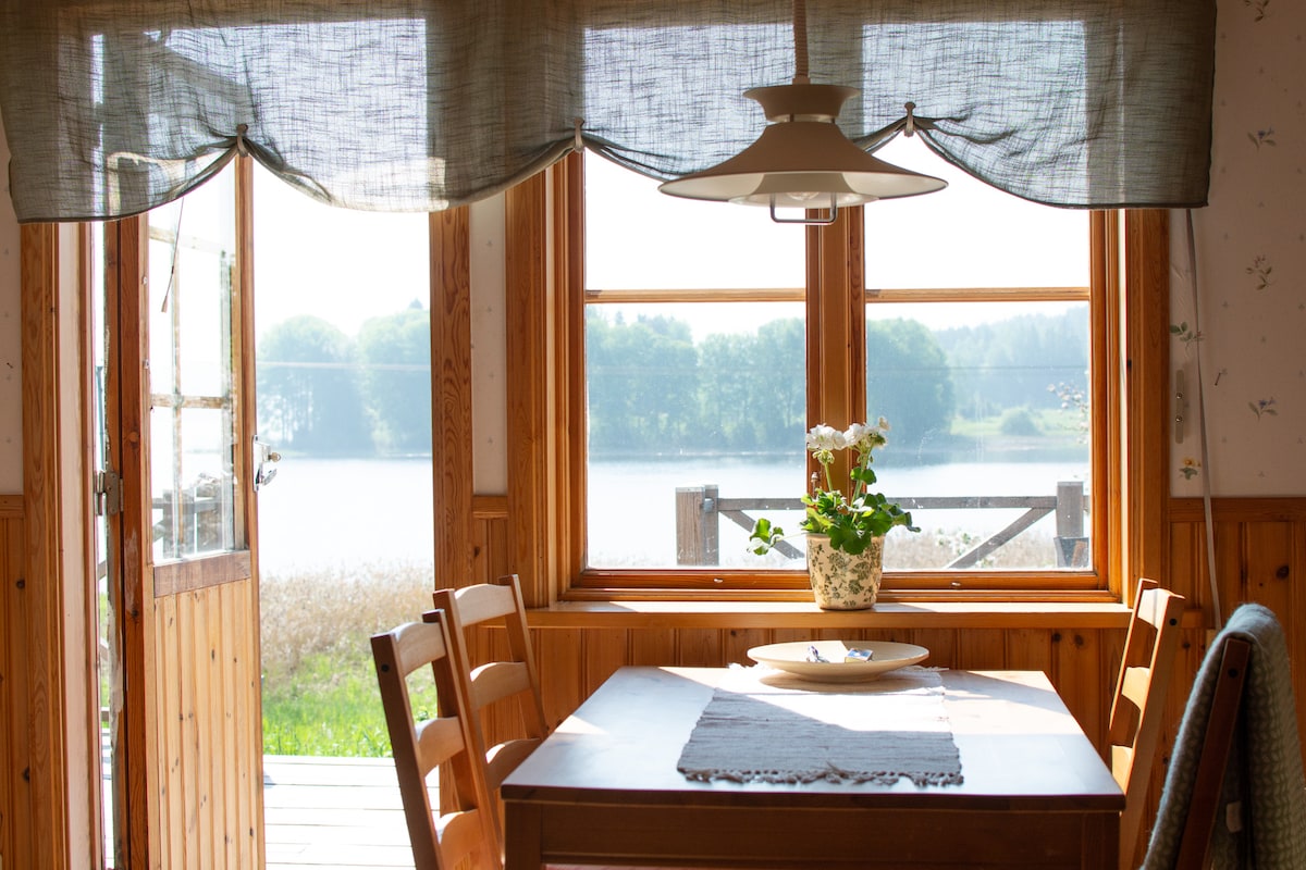 A dining area features a wooden table surrounded by chairs, positioned near large windows. Natural light floods the space, highlighting the floral centerpiece. Soft fabric curtains frame the windows, offering a view of the serene lake and surrounding greenery.