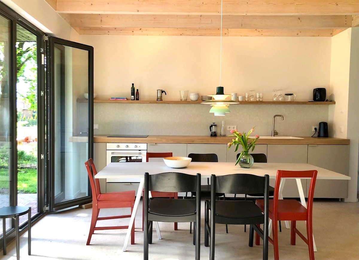 A modern kitchen and dining area features sleek cabinetry and a spacious dining table surrounded by colorful chairs. Large glass doors open to the garden, allowing natural light to illuminate the space. Kitchen appliances and a countertop are neatly arranged for convenience.