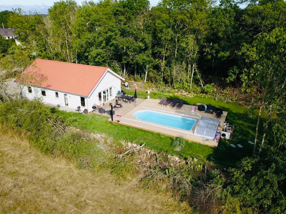An aerial view of a single-story house set amidst greenery. A rectangular pool is surrounded by a wooden deck with sun loungers. The house is framed by trees, offering a sense of seclusion, with outdoor seating visible near the pool.