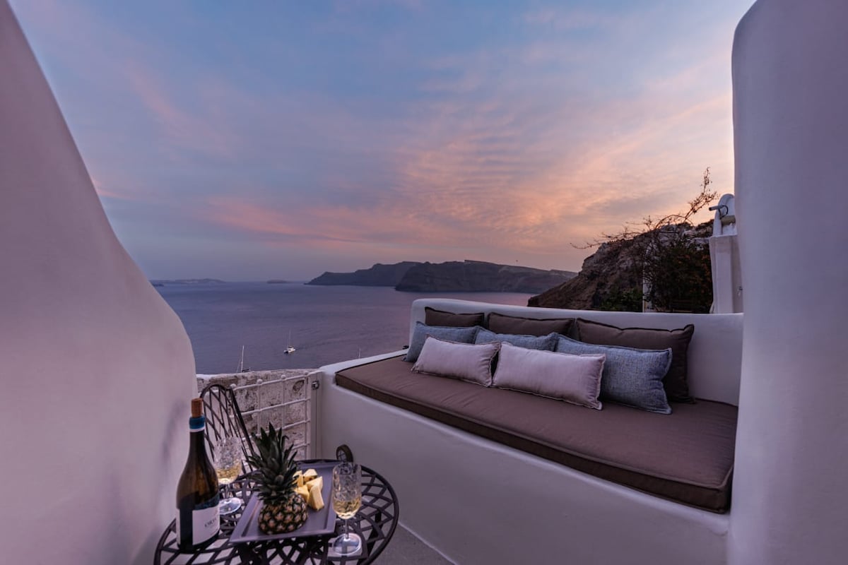 A private terrace is presented with a cozy seating area, featuring plush cushions in neutral tones. A small table holds a bottle of wine and two glasses, alongside a pineapple. The backdrop showcases a tranquil sea view during sunset, with soft hues in the sky.