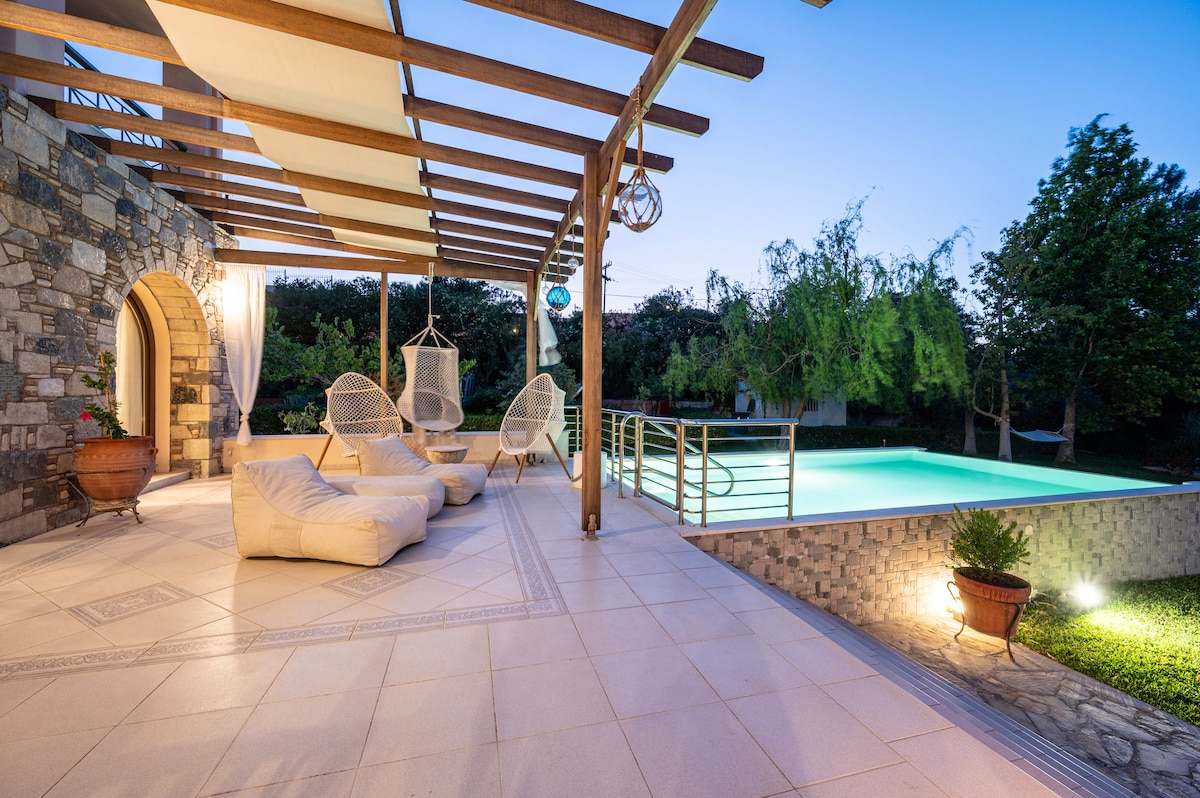 A spacious outdoor terrace features comfortable lounge seating, including a large bean bag and hanging chairs. The private pool glows softly in the evening light, surrounded by lush greenery. The stone walls complement the modern design, creating a serene atmosphere for relaxation.