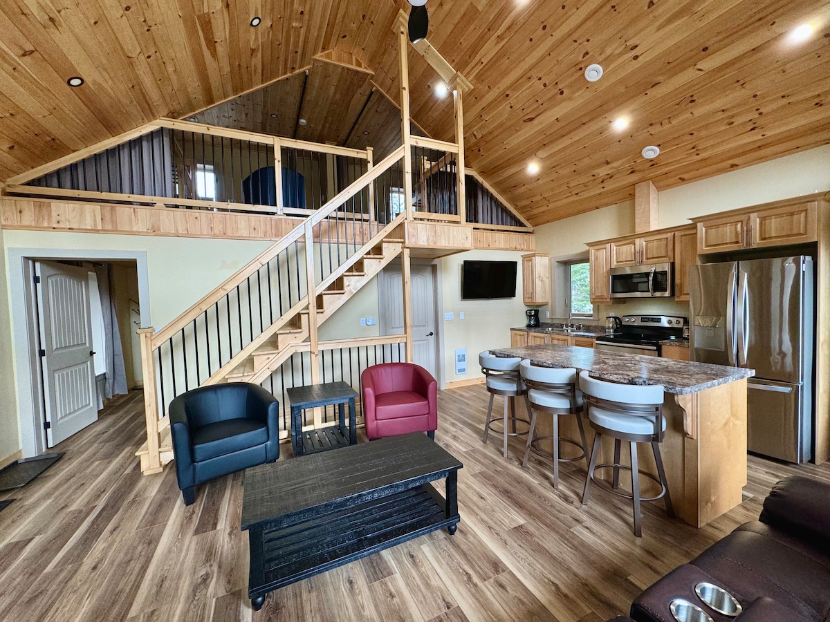 An open living space features a wooden staircase leading to a loft area. The main floor includes a kitchen with modern appliances and a dining area with a dark coffee table and light-colored bar stools. Comfortable seating is arranged with two armchairs in contrasting colors.