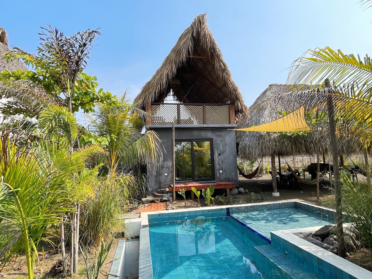 A thatched-roof casita is surrounded by tropical greenery, featuring a private pool with clear blue water. Hammocks are positioned nearby under shaded areas, inviting relaxation. The building’s exterior combines natural materials and large windows, creating a connection to the outdoors.