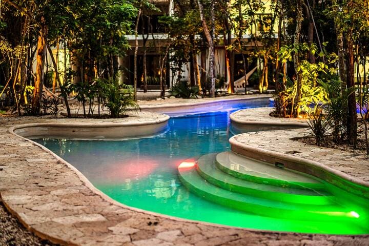 A winding pool is surrounded by lush tropical vegetation, featuring illuminated steps that lead into the water. Soft lighting in shades of blue and green reflects off the surface, creating a serene atmosphere that invites relaxation.