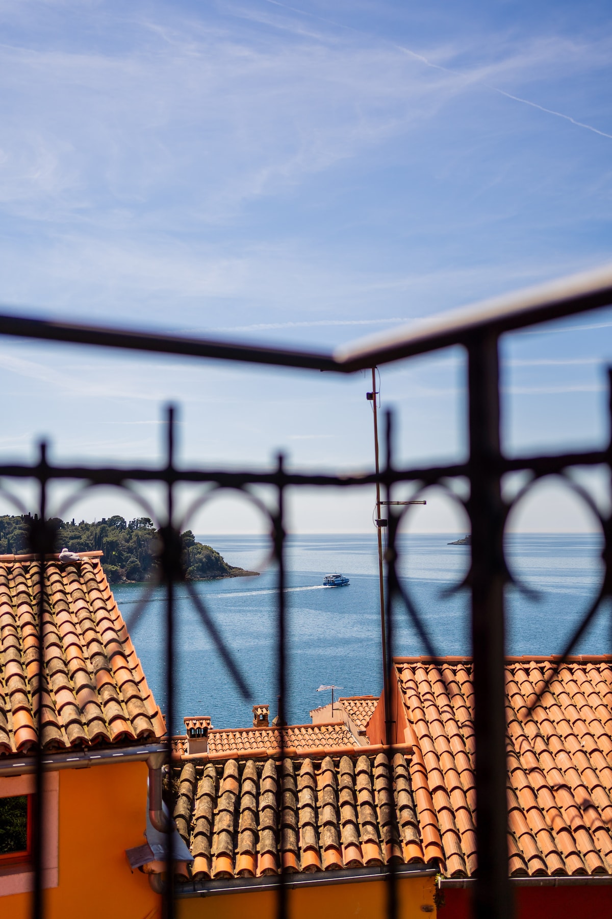 A view from a balcony showcases a serene coastal scene, with a sparkling blue sea extending to the horizon. The rooftops of vibrant buildings are framed by decorative metalwork, providing a glimpse of the picturesque surroundings under a clear sky.