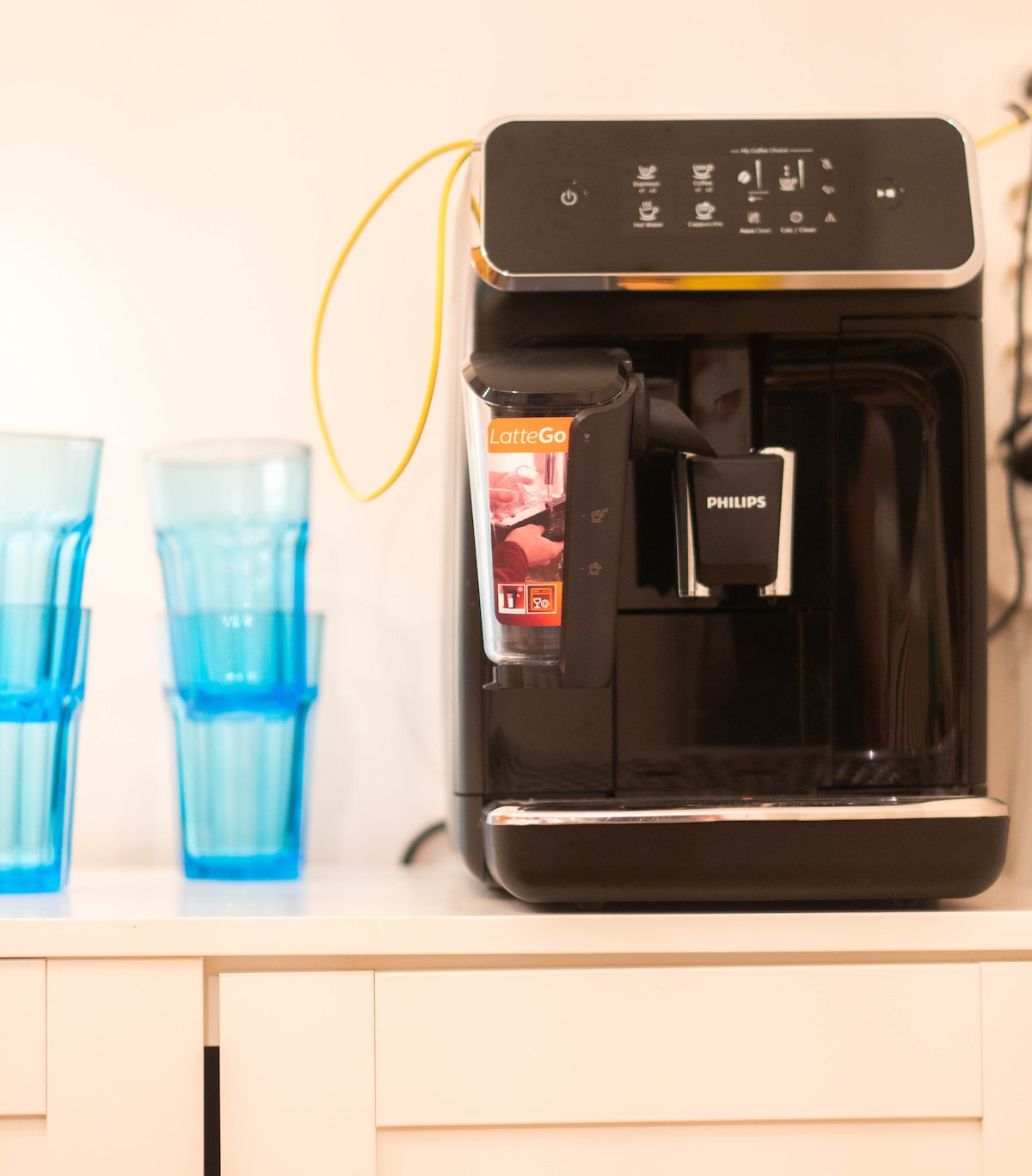 A modern coffee machine is positioned on a countertop, featuring a sleek black design and user-friendly control panel. Next to it, several clear blue drinking glasses are arranged, adding a pop of color to the kitchen space.
