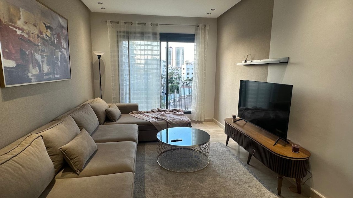 A spacious living area is highlighted by a large, L-shaped beige sofa positioned near a round glass coffee table. A flat-screen TV is mounted on a sleek shelf, while natural light filters through sheer curtains, illuminating the modern decor and an abstract artwork on the wall.