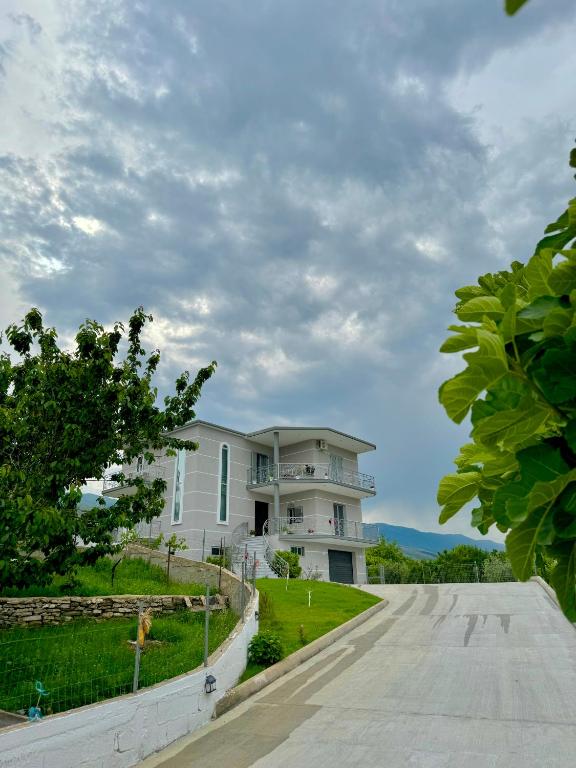 A modern three-story building is set against a backdrop of cloudy skies. Lush greenery surrounds the property, and a gently sloping driveway leads up to the entrance. Balconies are visible on the upper levels, offering views of the landscape.