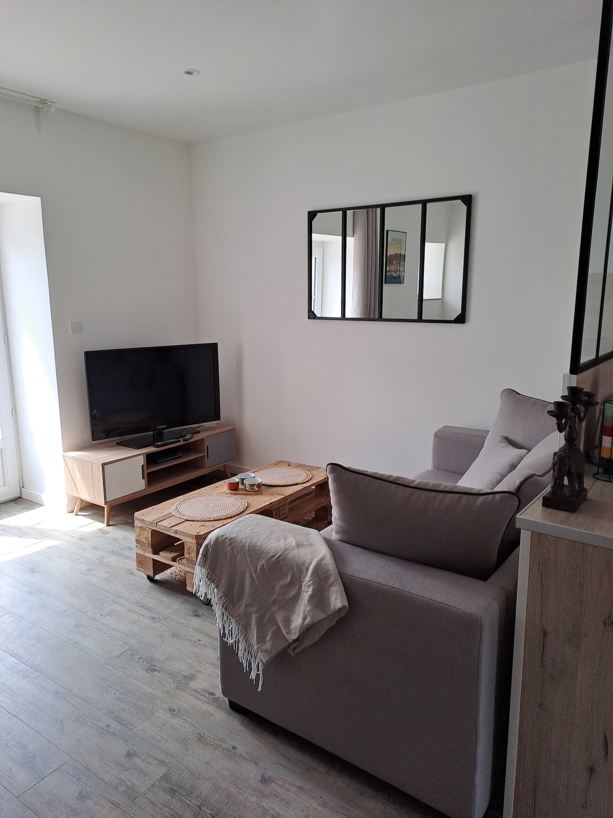A bright living area features a television on a wooden media console. A light-colored sofa is paired with a wooden pallet coffee table. Natural light fills the space through an adjoining door, with a wall mirror reflecting the room's ambiance.