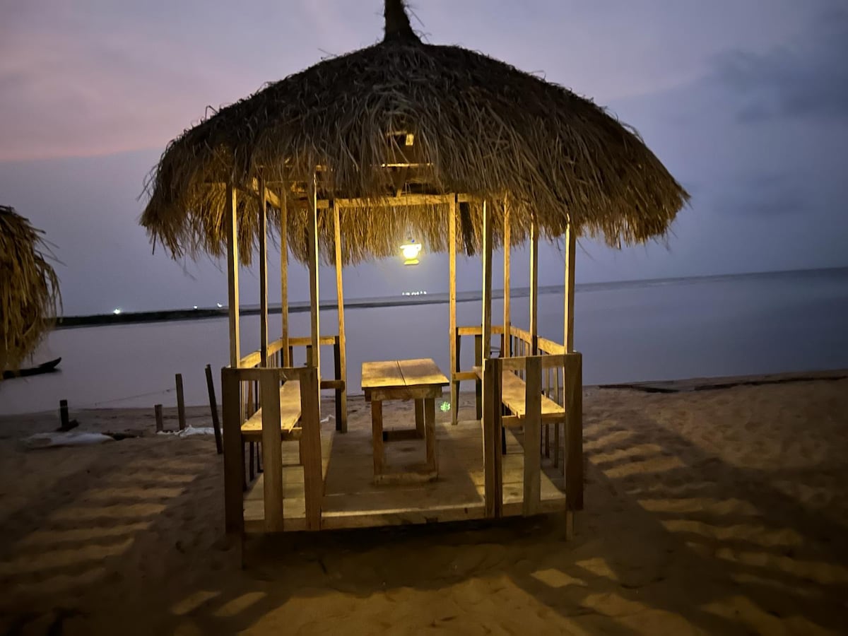 A coastal gazebo constructed of wood and adorned with a thatched roof is positioned on a sandy beach. Soft ambient lighting illuminates the structure, which features a central table surrounded by benches, facing the calm water.