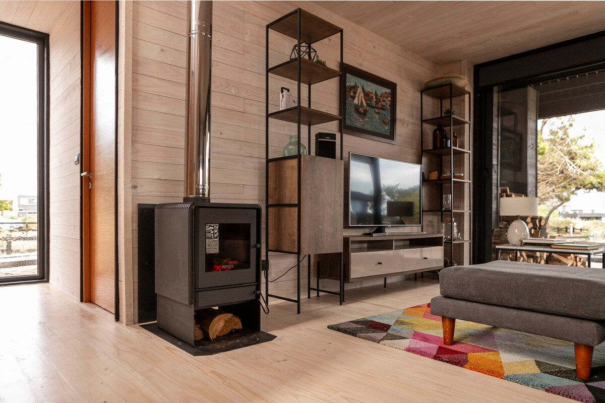 A modern living area features a wood-burning stove positioned beside a contemporary shelving unit. A television is mounted on a clean-lined console, while a large window offers natural light and views of the outdoors. A colorful area rug adds warmth to the light wood flooring.
