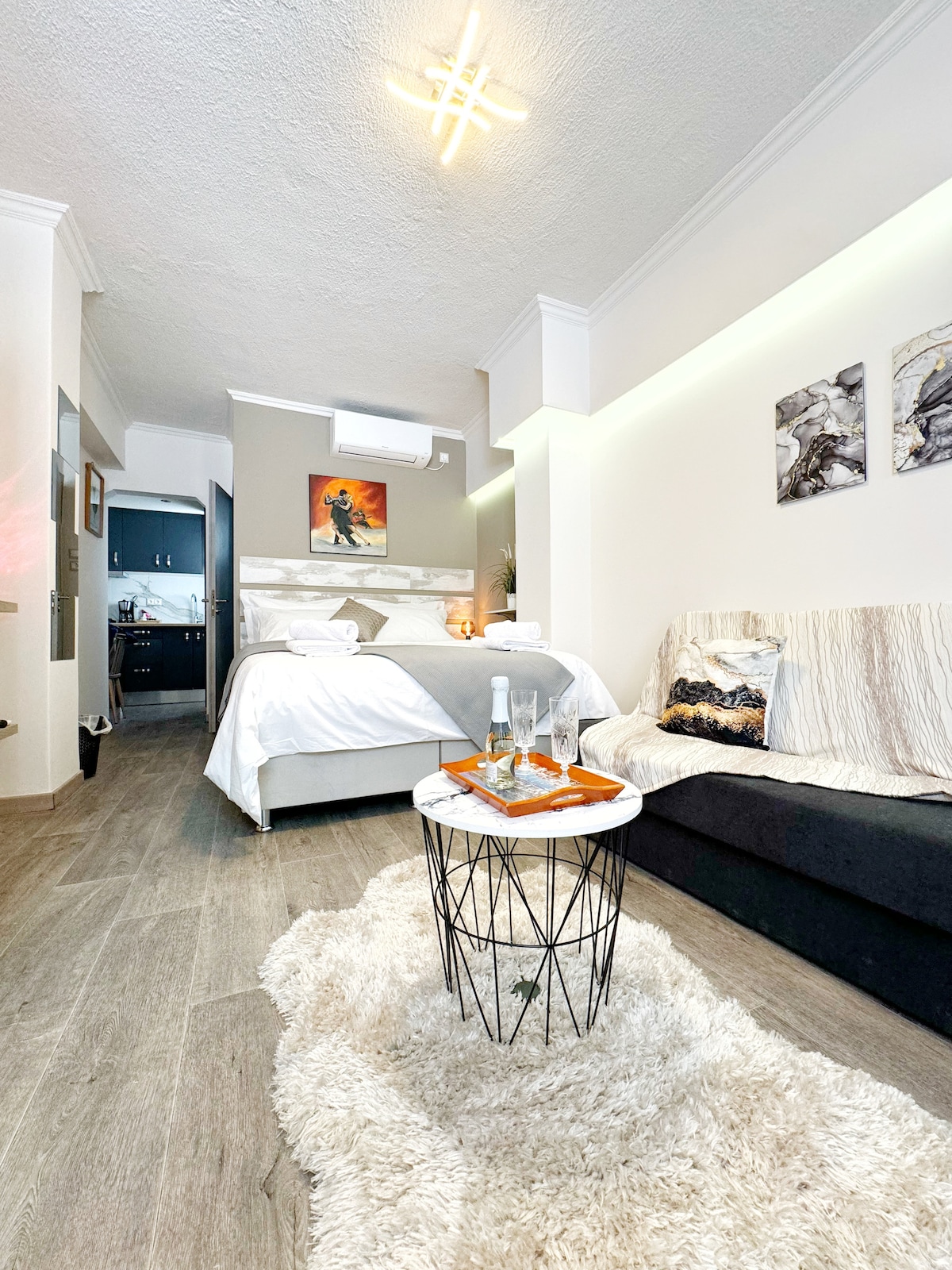 A well-lit living space features a spacious bed dressed in soft linens, complemented by neutral-toned decor. A cozy sofa is positioned beside a small circular table, which holds a bottle and glasses. Artwork and soft textures add inviting touches to the room.