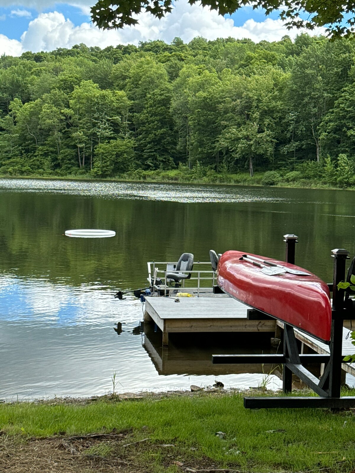 A red kayak is positioned on a floating dock, which extends toward a calm lake reflecting surrounding greenery. Soft ripples disturb the water's surface, while a lush forested area in the background provides a serene backdrop.