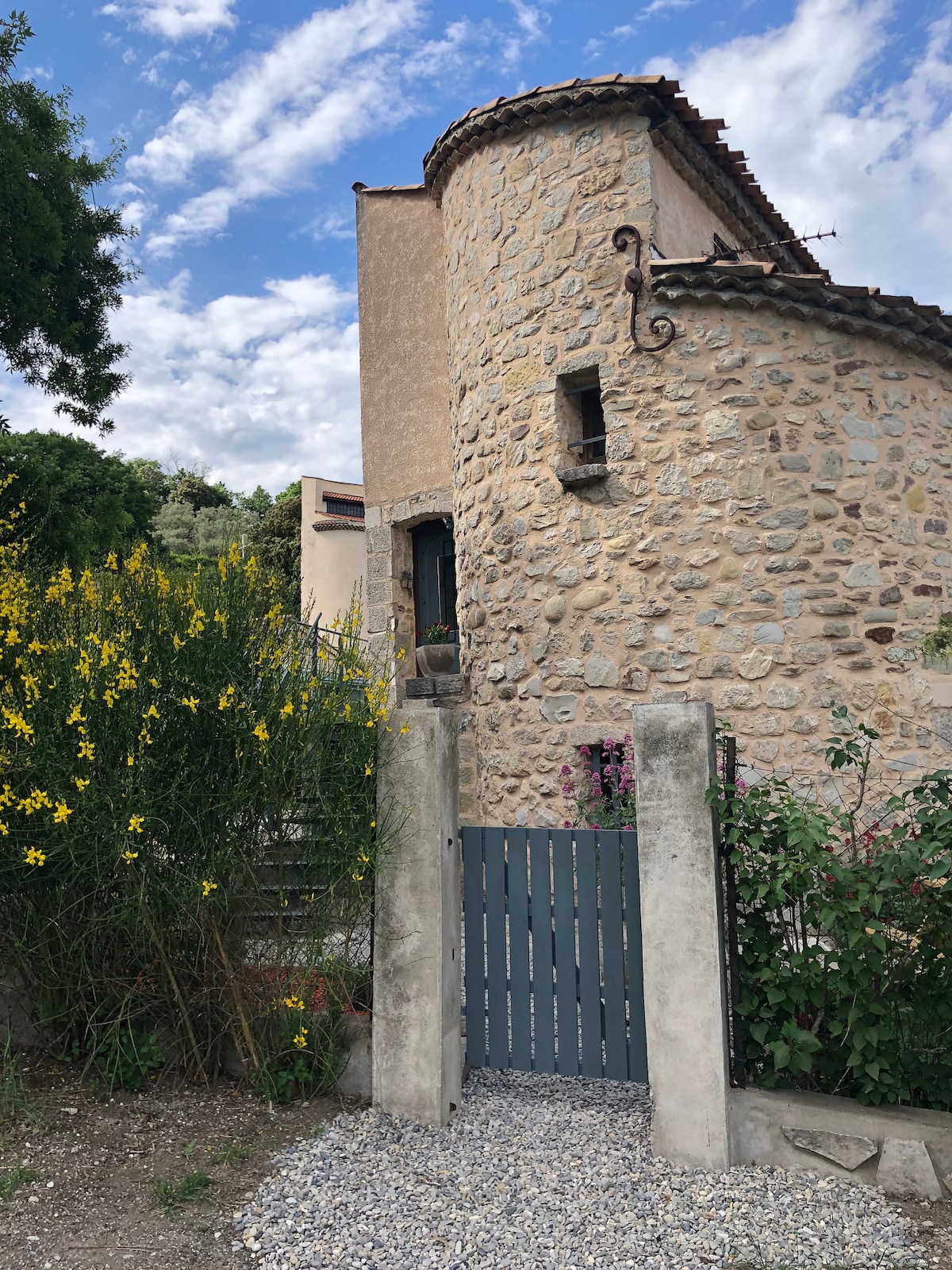 A stone structure, characterized by a round turret and a tiled roof, is set against a backdrop of a partly cloudy sky. Surrounding vegetation includes blooming yellow flowers near a gray gravel path leading to a wooden gate.
