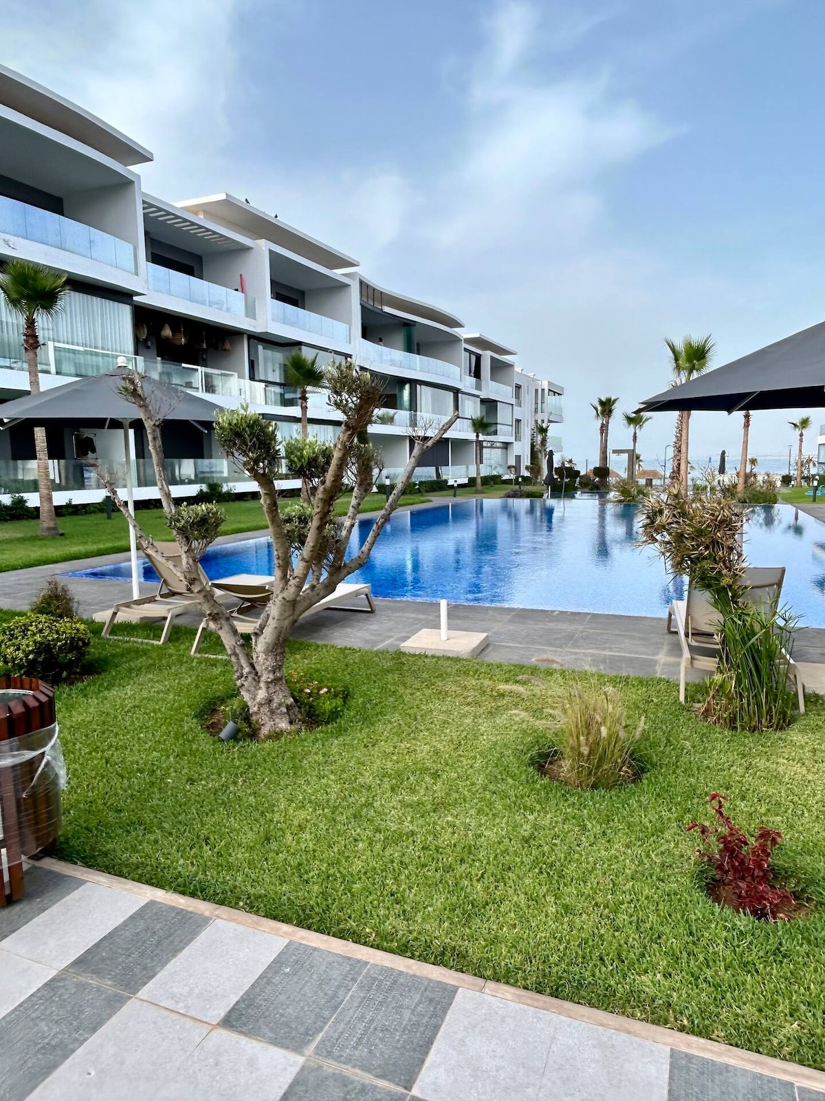 A serene outdoor area features a large swimming pool bordered by lush green grass and palm trees. Sun loungers are arranged under shaded umbrellas, offering a comfortable place for relaxation. Modern apartment buildings provide a backdrop, enhancing the tranquil atmosphere of the space.