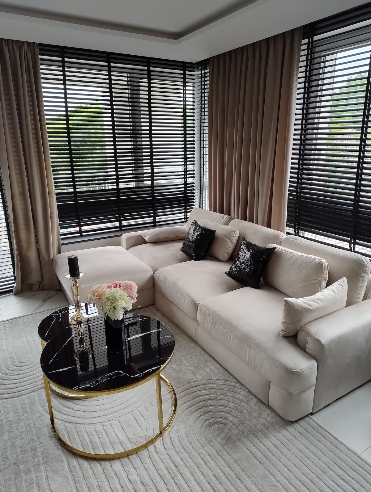 A spacious living area is furnished with a light-colored sectional sofa adorned with decorative black cushions. A striking black coffee table with a gold base is centered on a textured rug, complemented by elegant window treatments allowing filtered light to enhance the space.