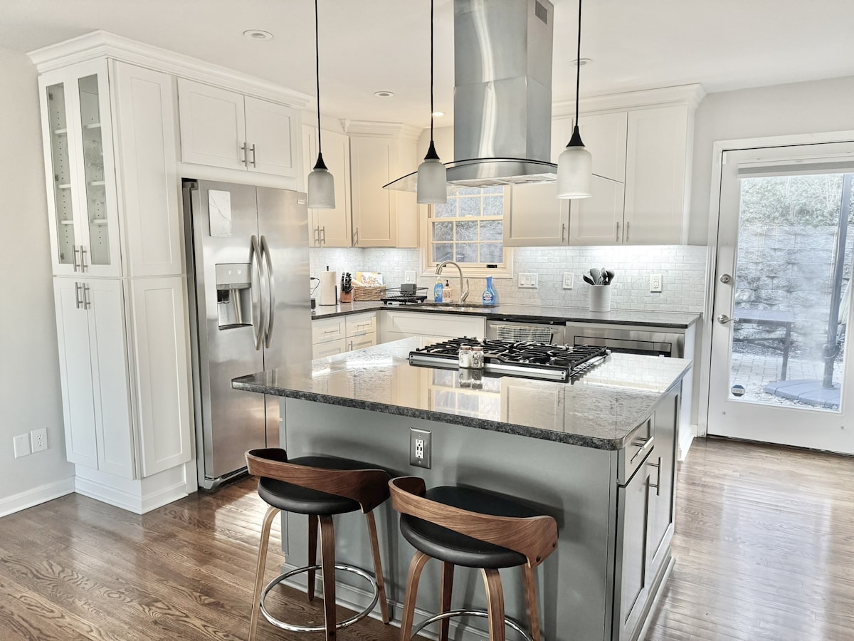 The modern kitchen features sleek cabinetry and a central island with seating for two. Stainless steel appliances, including a refrigerator and gas stove, are visible. Natural light enters through a nearby door, which leads to an outdoor space, enhancing the bright and open atmosphere.