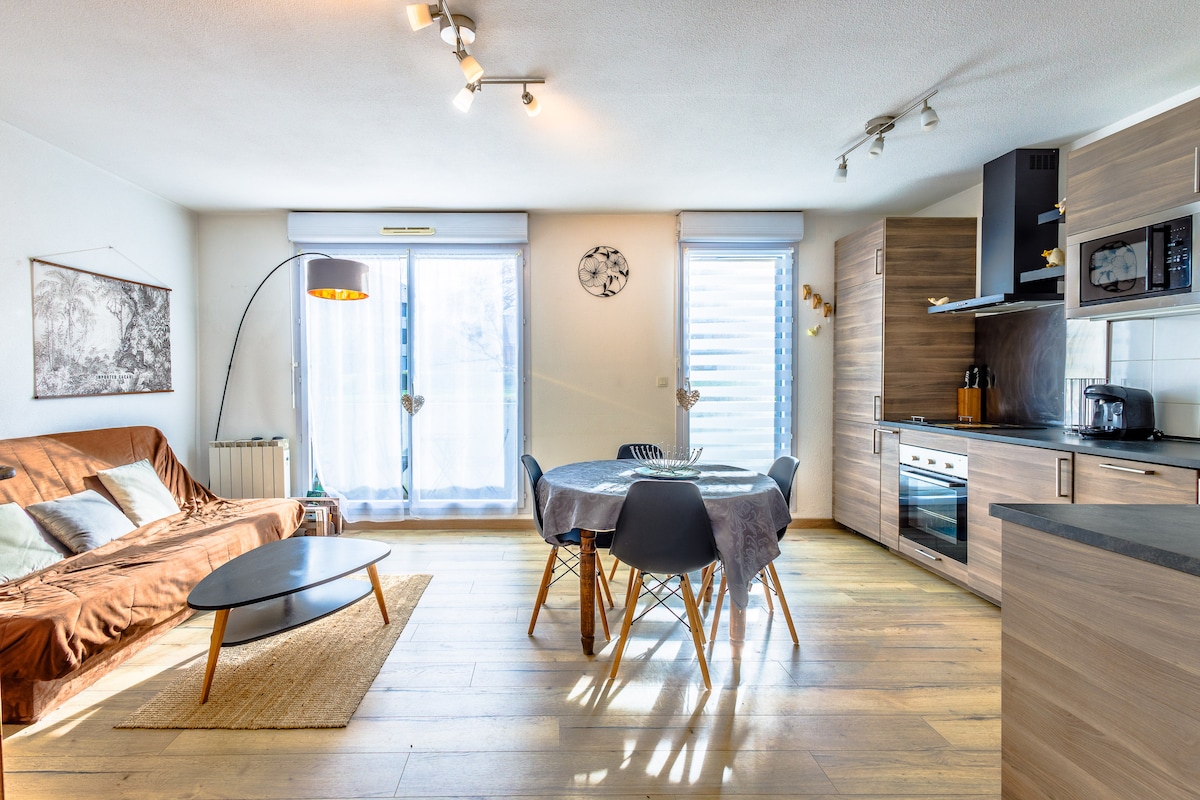 A spacious living area features a convertible sofa, a round dining table with chairs, and a modern kitchen with sleek cabinetry. Natural light floods through large windows adorned with light curtains, accentuating the warm wood flooring. A decorative light fixture enhances the contemporary design.