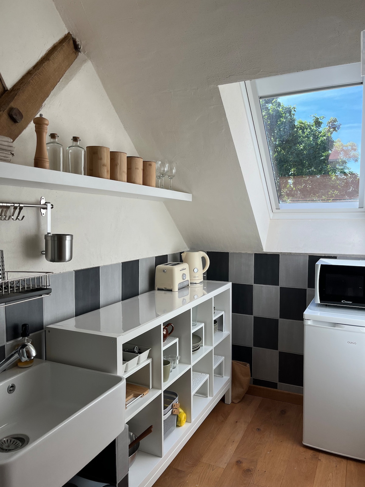 A modern kitchen area is displayed with sleek white cabinetry and black and white tiled walls. Natural light enters through a skylight, highlighting the organized shelves filled with various kitchen items. A compact refrigerator and a microwave are positioned for convenience.