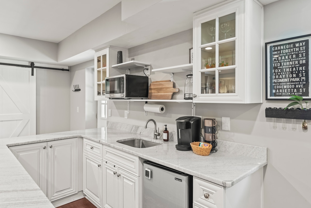 The kitchen area features white cabinetry with glass doors, offering a view of neatly arranged dishware. A grey marble countertop provides space for meal preparation. Essential appliances include a microwave, coffee maker, and compact refrigerator, ensuring convenience for guests during their stay.
