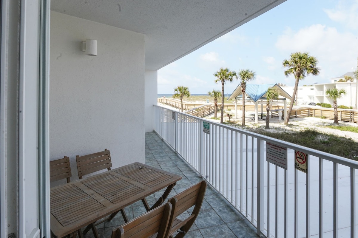A covered patio area features a wooden dining table accompanied by several matching chairs. The railing provides an unobstructed view of palm trees and a nearby boardwalk leading to the beach, under a bright sky with clouds.