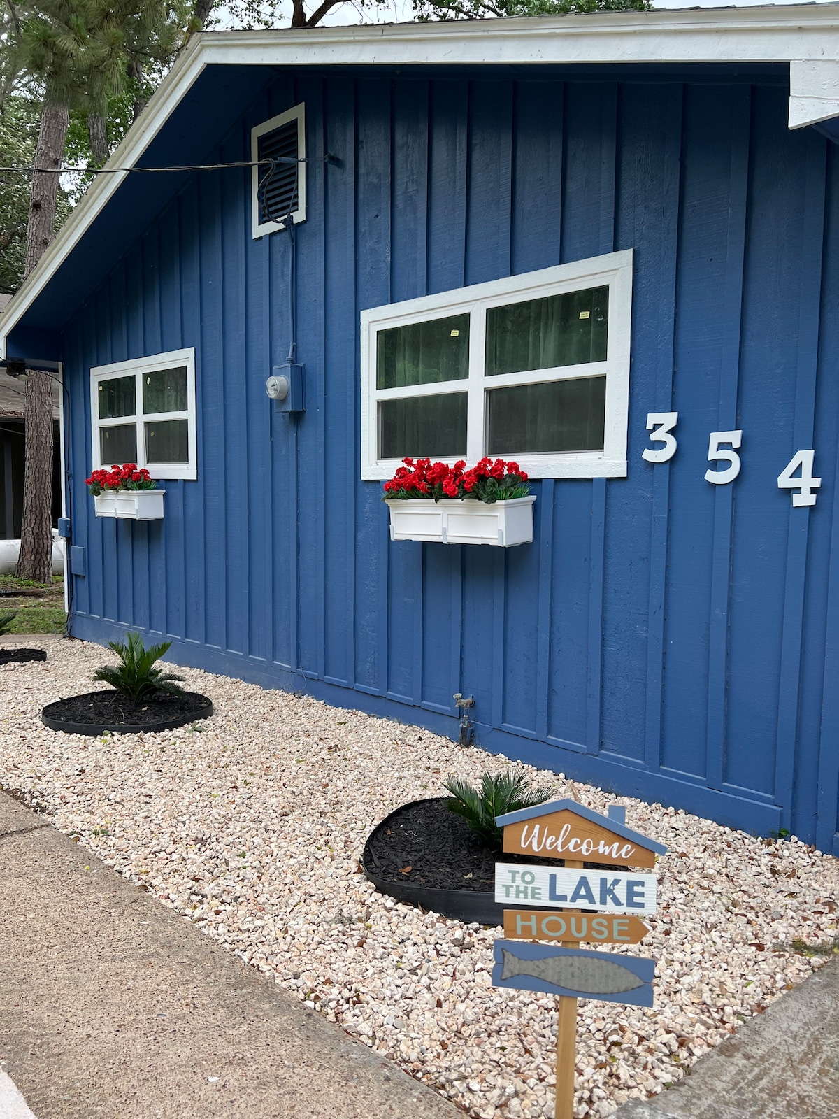 A charming blue cabin exterior is depicted, featuring three white-framed windows adorned with red flowers. Stone landscaping is positioned around the base, while a welcome sign is placed on the pathway, indicating the entrance to the lake house.