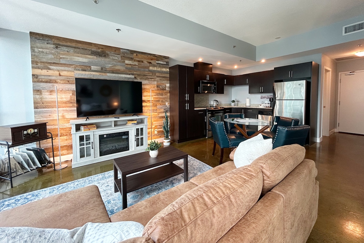 A spacious living area is highlighted by a large flat-screen TV mounted on a rustic wood wall. The modern kitchen features dark cabinetry and a dining area with a round table and stylish chairs. A cozy sofa and decorative accents create a welcoming atmosphere.