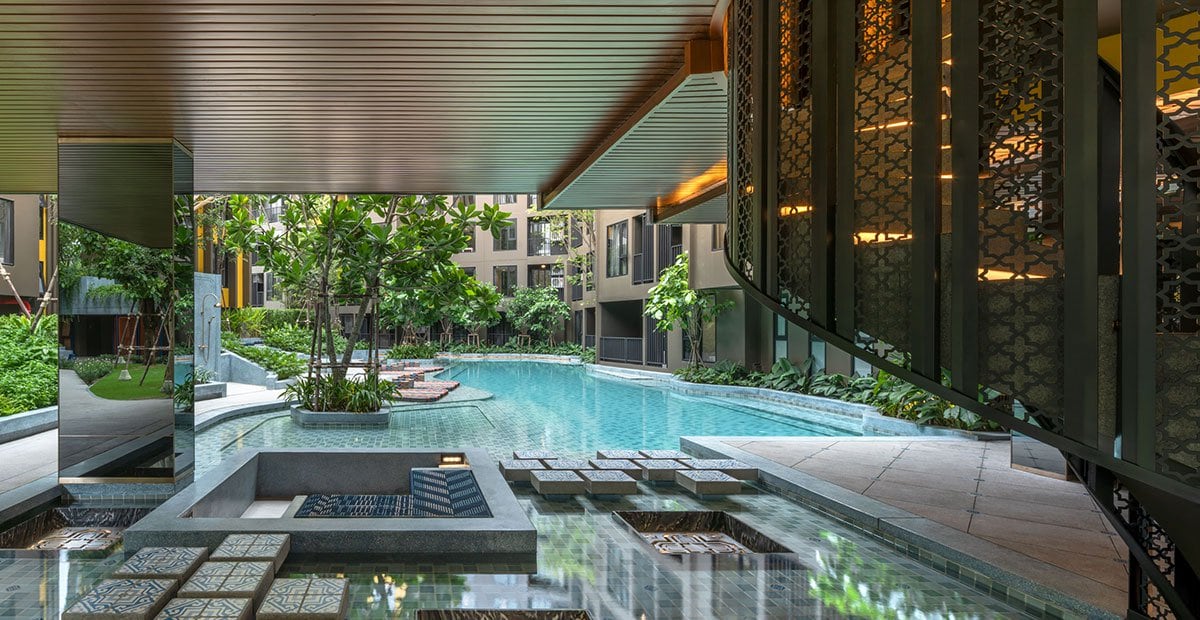 The image displays a serene pool area surrounded by lush greenery, with a reflective water surface. Comfortable seating is arranged along the edge, and trees provide shade. Architectural details, including patterned screens and a modern staircase, contribute to a tranquil ambiance.