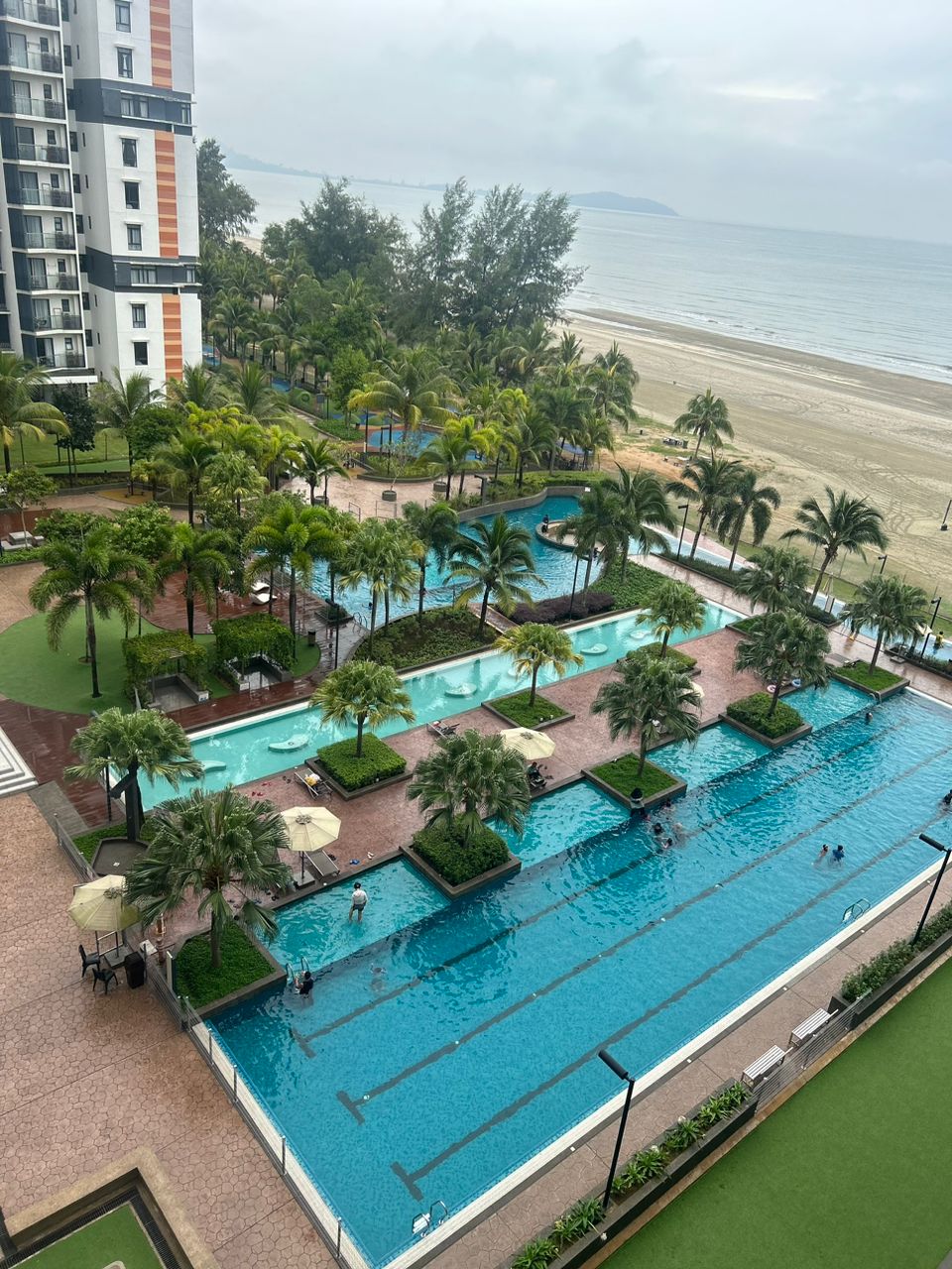 An aerial view reveals a landscaped pool area surrounded by palm trees. Multiple swimming pools are set among pathways and lounge chairs, leading to a sandy beach and the ocean in the background, creating a serene atmosphere.