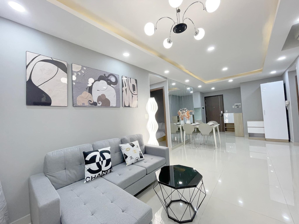 A modern living area is depicted, featuring a grey sofa adorned with decorative pillows. A geometric coffee table sits in front of the sofa. The space is illuminated by a stylish light fixture, with artwork displayed on the walls and an open dining area visible in the background.