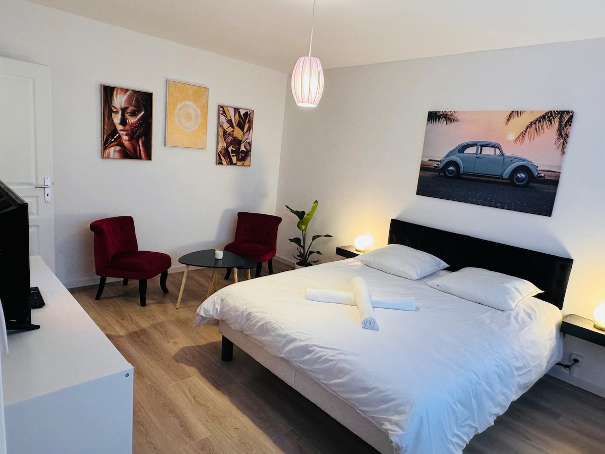 A well-lit sleeping area features a spacious bed with soft linens and decorative pillows. Two red chairs are positioned near a small table, complemented by wall art showcasing vibrant designs. A television is visible across from the bed, with ambient lighting enhancing the room's features.