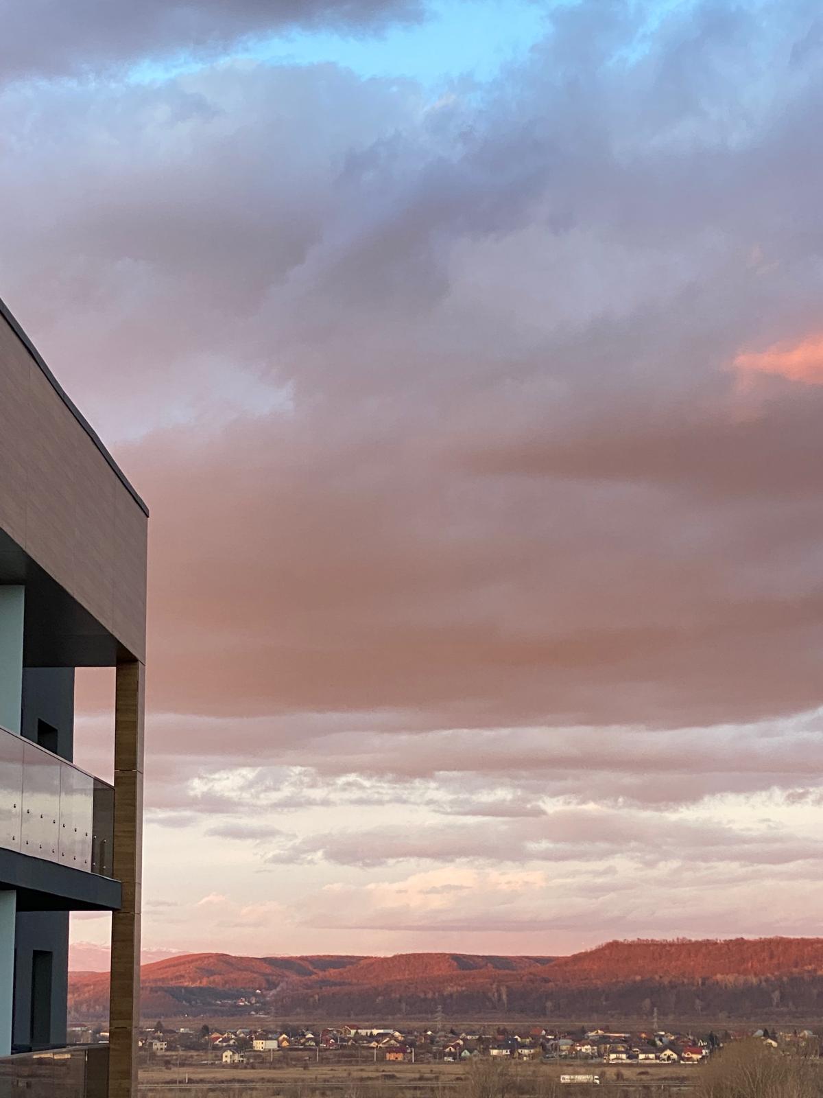 A view from the apartment features a soft gradient of pastel pink and blue hues in the sky, with wispy clouds reflecting warm light. The modern architectural shape of the building is visible in the foreground, providing a contemporary contrast to the natural scenery.