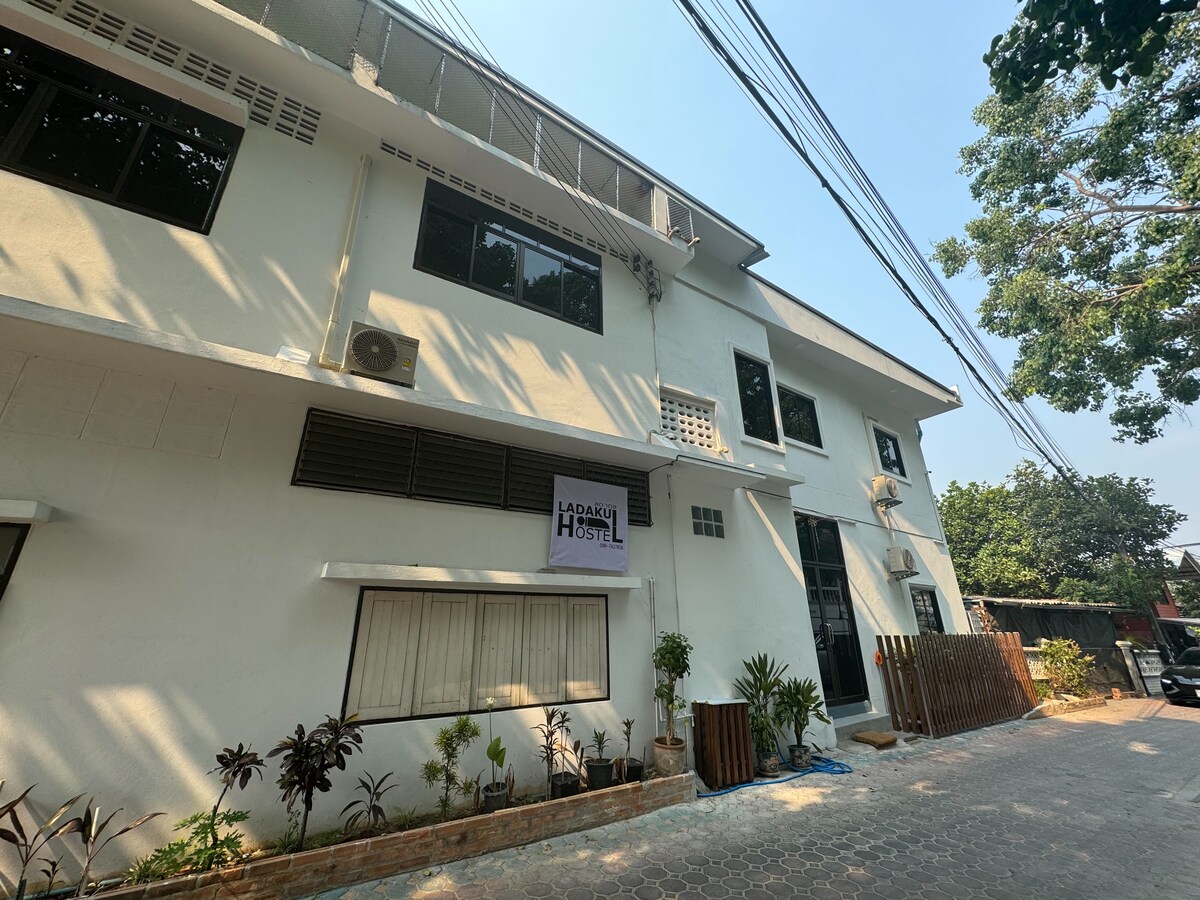 The building facade is characterized by a clean white exterior and multiple windows allowing natural light to enter. A sign indicating the hostel name is prominently displayed. Surrounding greenery enhances the welcoming atmosphere. A paved pathway leads to the entrance.