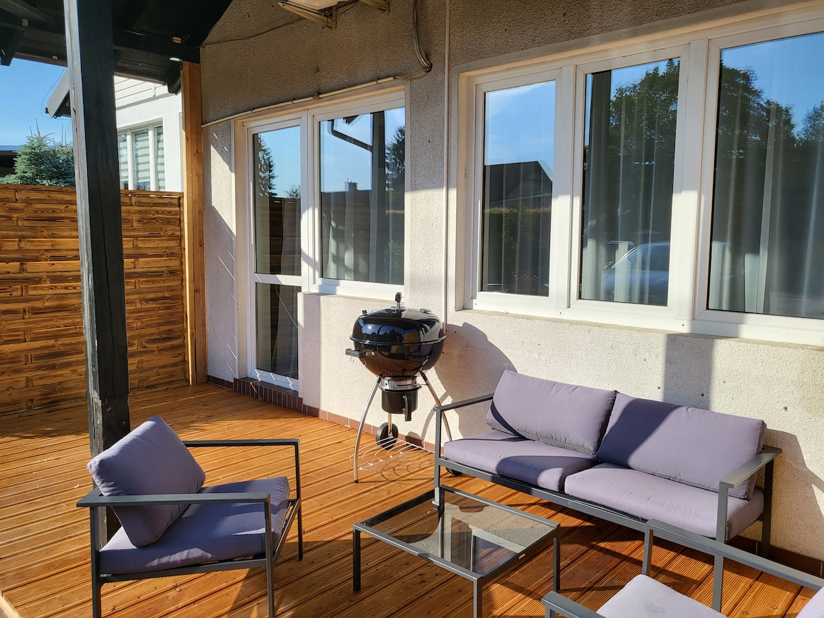 An outdoor terrace is shown, featuring a wooden deck and comfortable seating arrangements. A round charcoal grill is positioned nearby, along with a glass-top table. Light filters through large windows, providing an airy atmosphere in this inviting space.