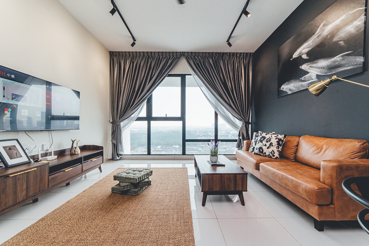The spacious living area features a large window with city views, framed by elegant curtains. A comfortable brown sofa is positioned beside a wooden coffee table. A smart TV is mounted on the wall, and decorative accents enhance the modern design.