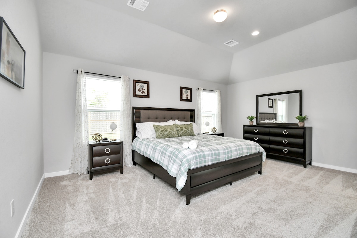 A spacious bedroom features a dark wood bed frame with a plaid comforter on a generous bed. Two windows allow natural light, complemented by sheer curtains. A dresser and mirror enhance the decor, while the plush carpet adds comfort. Green accents provide a calming touch.