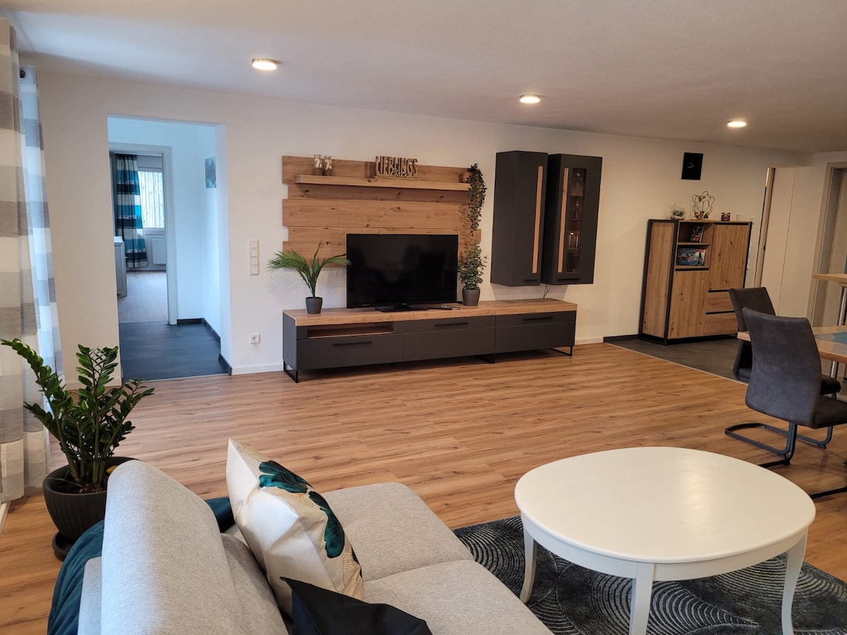 A spacious living area is featured with wooden flooring and neutral walls. A large television is mounted on a decorative wooden panel. A comfortable gray sofa, complemented by decorative cushions, is positioned near a round coffee table, with a plant nearby adding a touch of greenery.