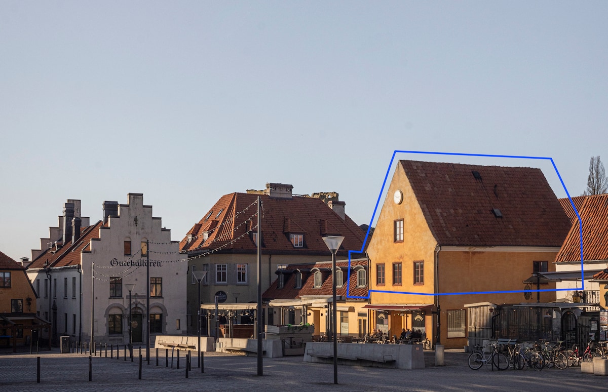A two-story yellow building with a sloped roof is positioned prominently at Stora Torget. Surrounding structures reflect a mix of historical and modern architecture. Clear skies are visible above, and bicycles are parked nearby, providing a sense of community in this vibrant square.