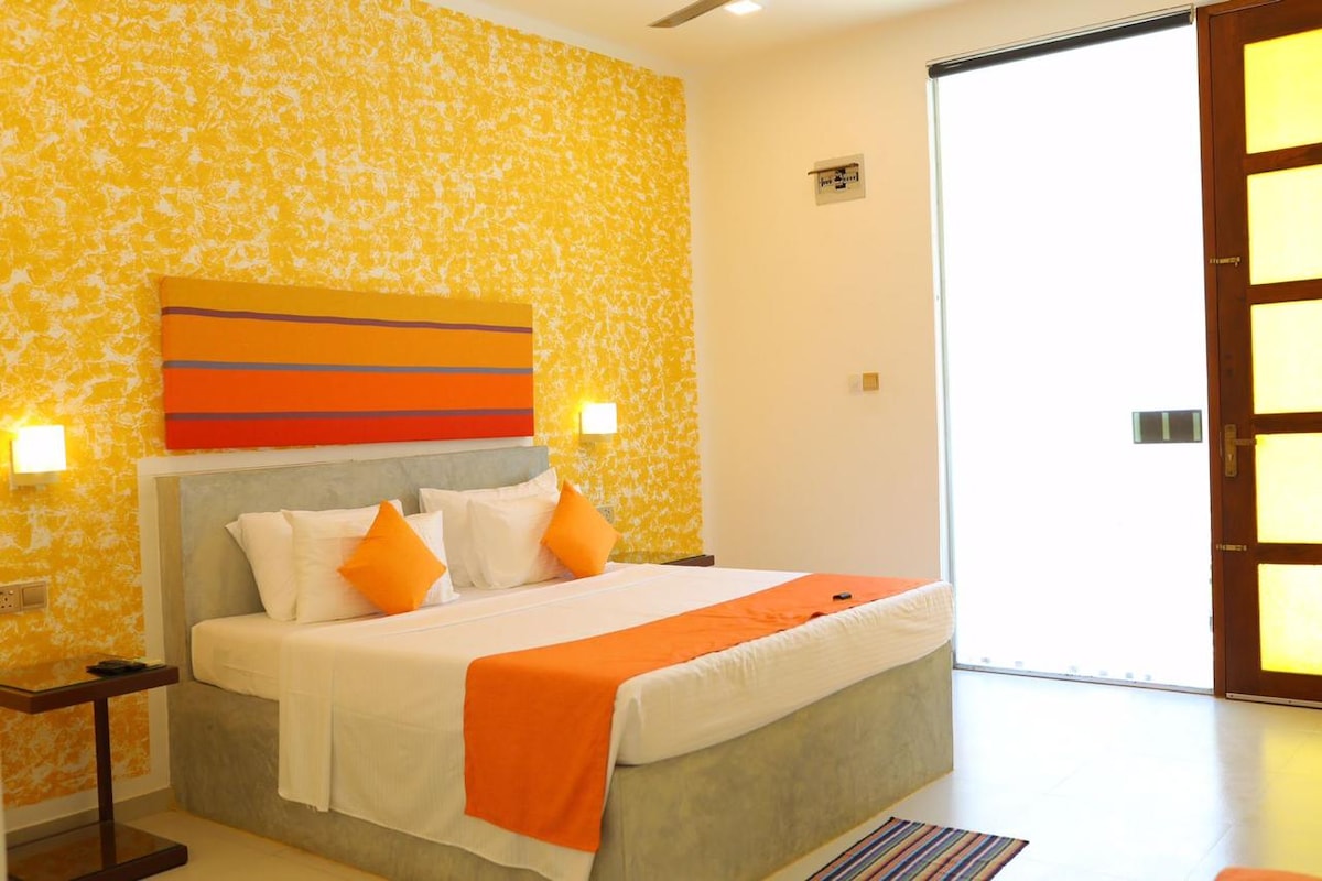 A well-lit room features a spacious bed with an orange and white color scheme. Two orange accent pillows sit against a textured yellow wall. A colorful striped rug is placed on the light-colored floor, and large glass doors allow natural light to enter the space.