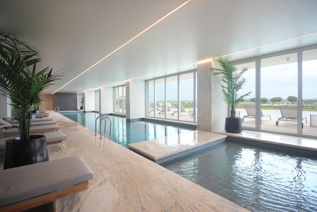 An indoor pool extends along a sleek marble surface, bathed in natural light from large floor-to-ceiling windows. Comfortable loungers line the area, with indoor plants adding a touch of greenery. Views of the outdoor landscape are visible through the glass panes.