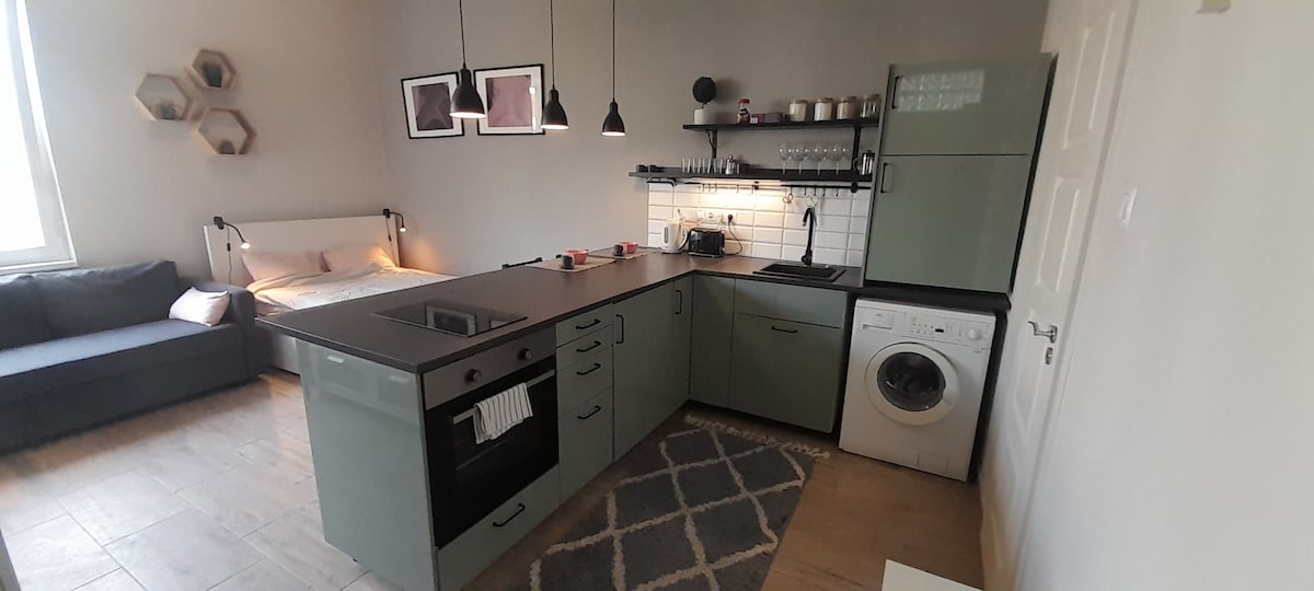 A modern kitchen area is paired with a living space, featuring an island counter with a built-in stove. Appliances include a sleek refrigerator and a washing machine. Soft lighting from pendant fixtures creates a warm ambiance, while geometric decor accents the walls.