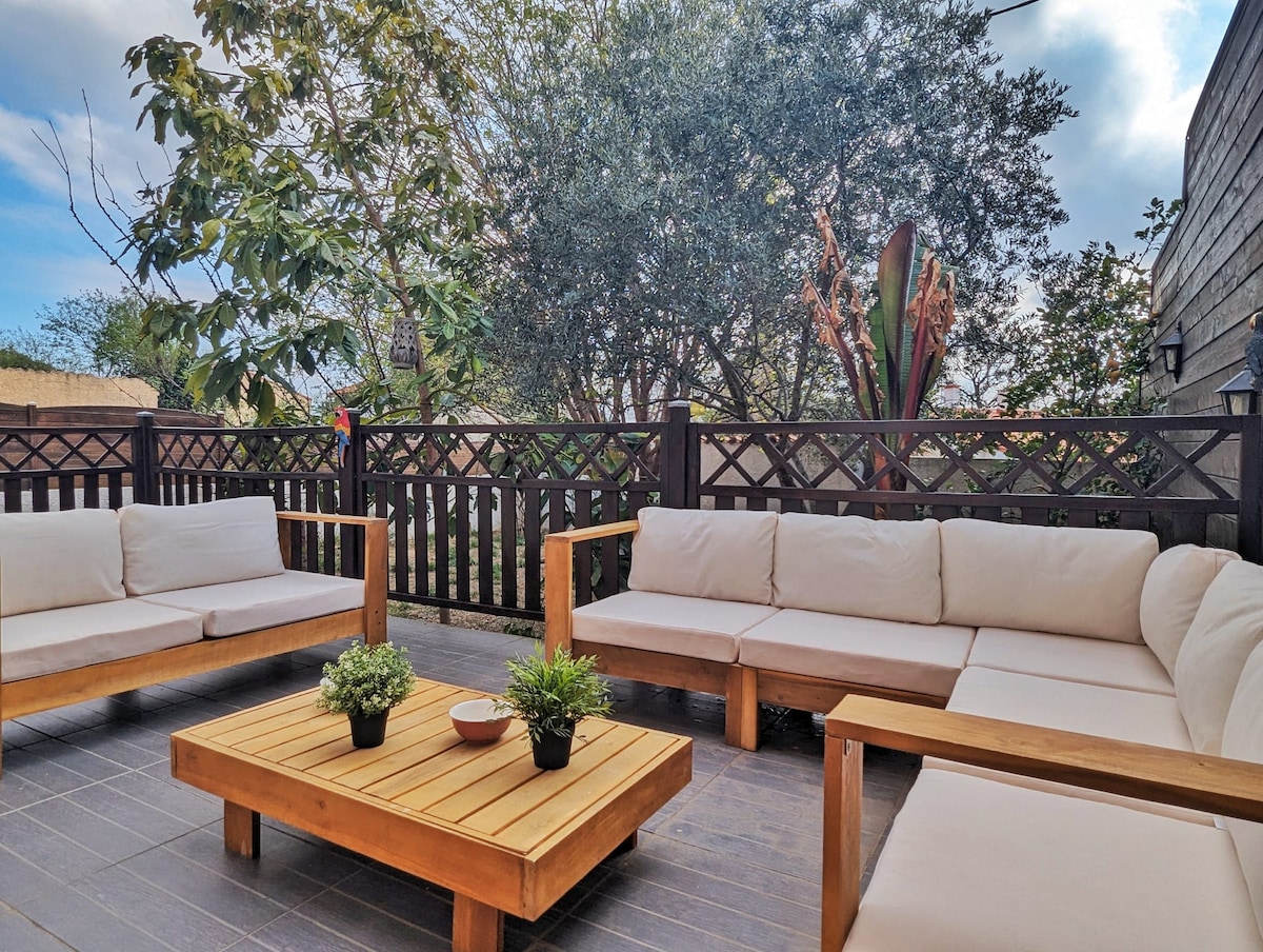 A spacious terrace is furnished with multiple light-colored seating areas, offering a relaxed atmosphere. A wooden coffee table sits in the center, accented by small potted plants. Surrounding greenery adds a natural touch to the space, enhancing the overall inviting environment.