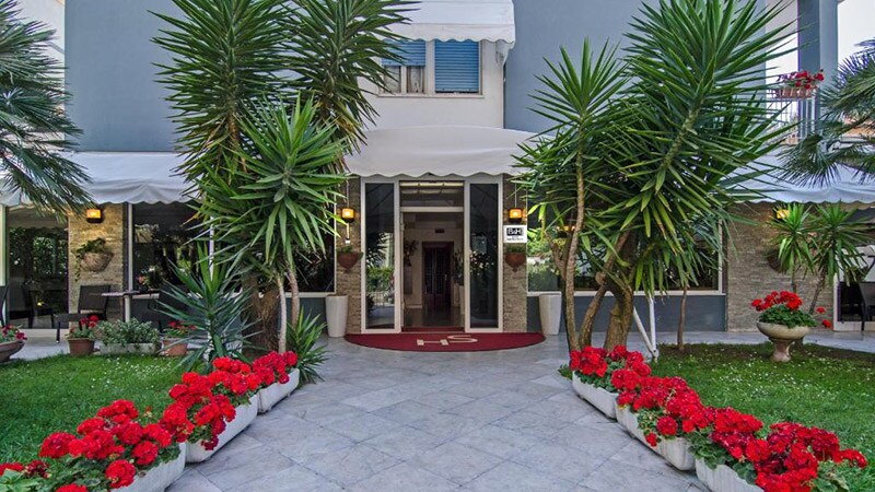 The entrance is framed by lush greenery and vibrant red flowers in planters. A welcoming path of stone tiles leads to the covered entryway, where large glass doors provide access to the interior. Palm trees add to the inviting ambiance of the exterior.