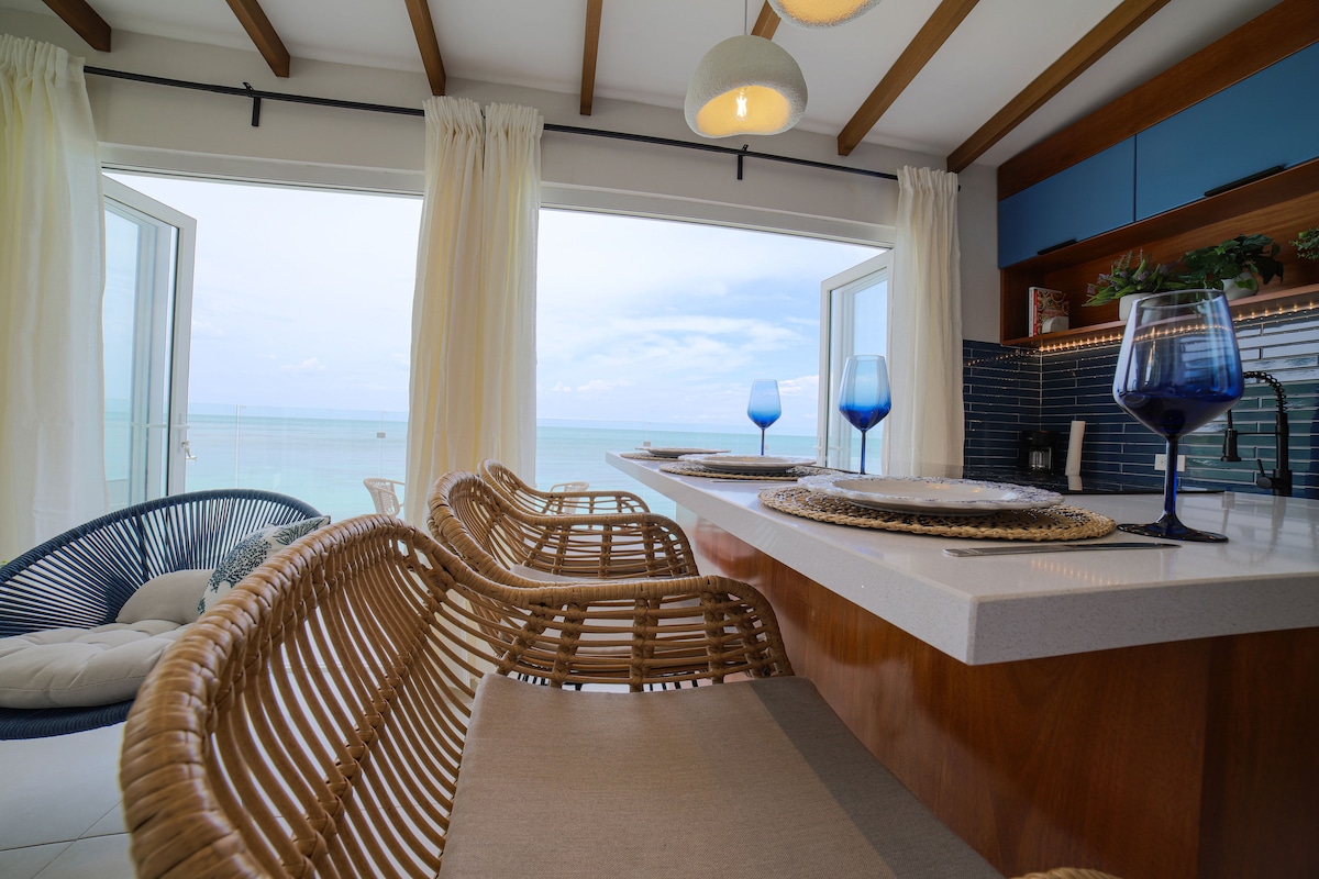 A bright kitchen area features a countertop with woven bar stools, overlooking a stunning ocean view. Floor-to-ceiling windows invite natural light, while soft curtains frame the scene. Glasses in blue tones are arranged on the counter, complementing the coastal decor.