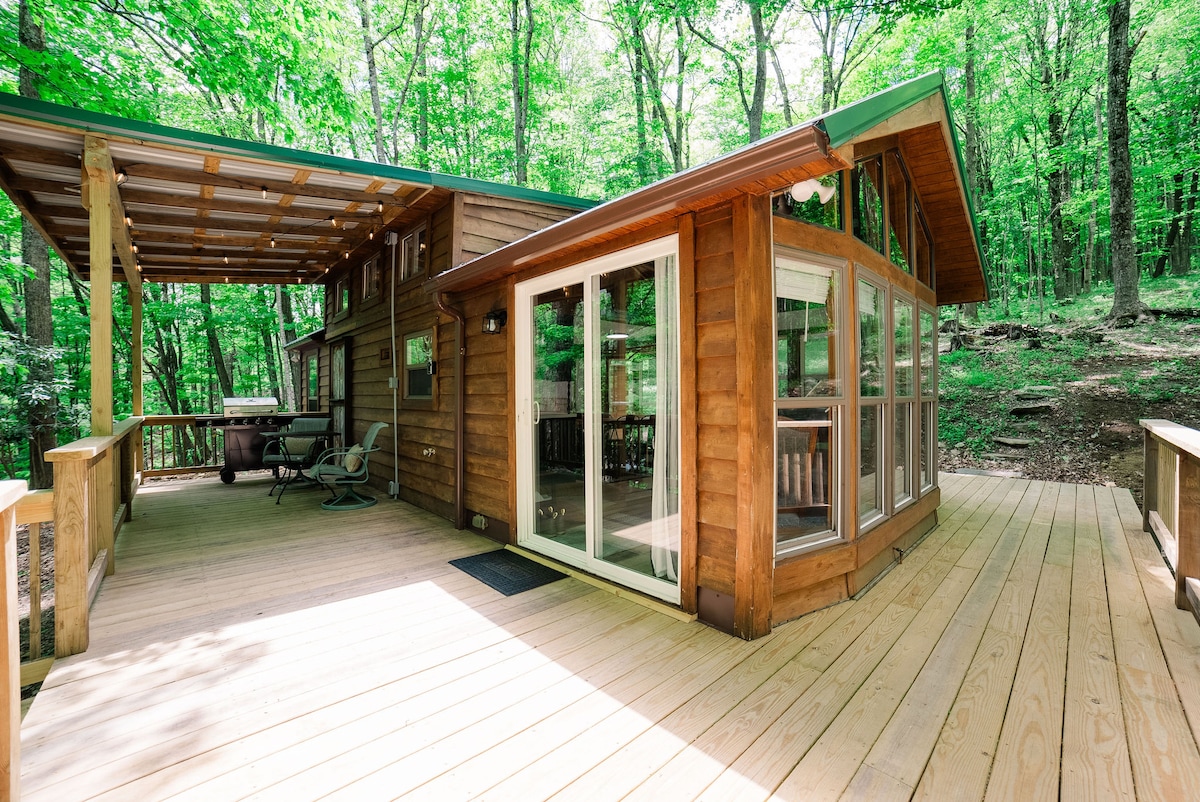 A spacious wood deck is presented, surrounded by lush green trees. Large glass doors provide a view of the interior, while a shaded area with seating can be seen. Natural light floods the deck, reflecting off the warm wood tones of the cabin structure.