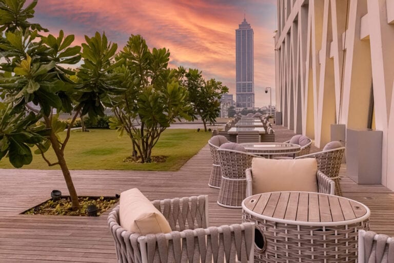 An outdoor terrace features woven seating arranged around circular tables. Green landscaping and plants provide a natural touch, while a city skyline is visible in the background, highlighted by a colorful sunset sky.